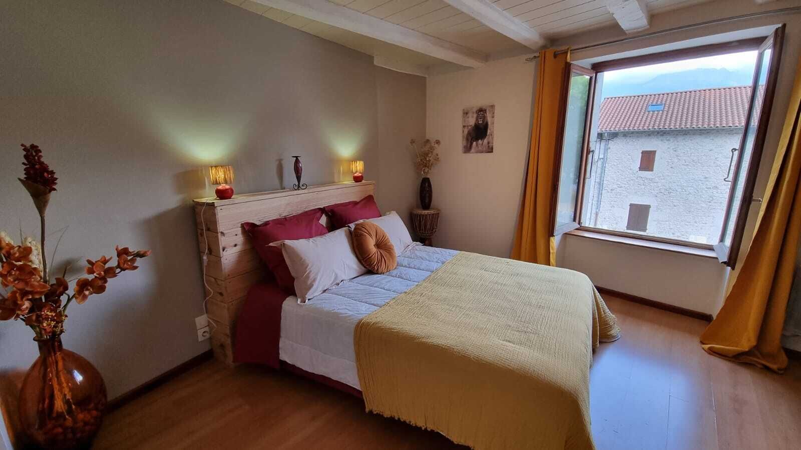 Un dormitori amb un llit, una tauleta de nit, un vas amb flors, una finestra amb cortines i vistes a edificis a l'exterior.