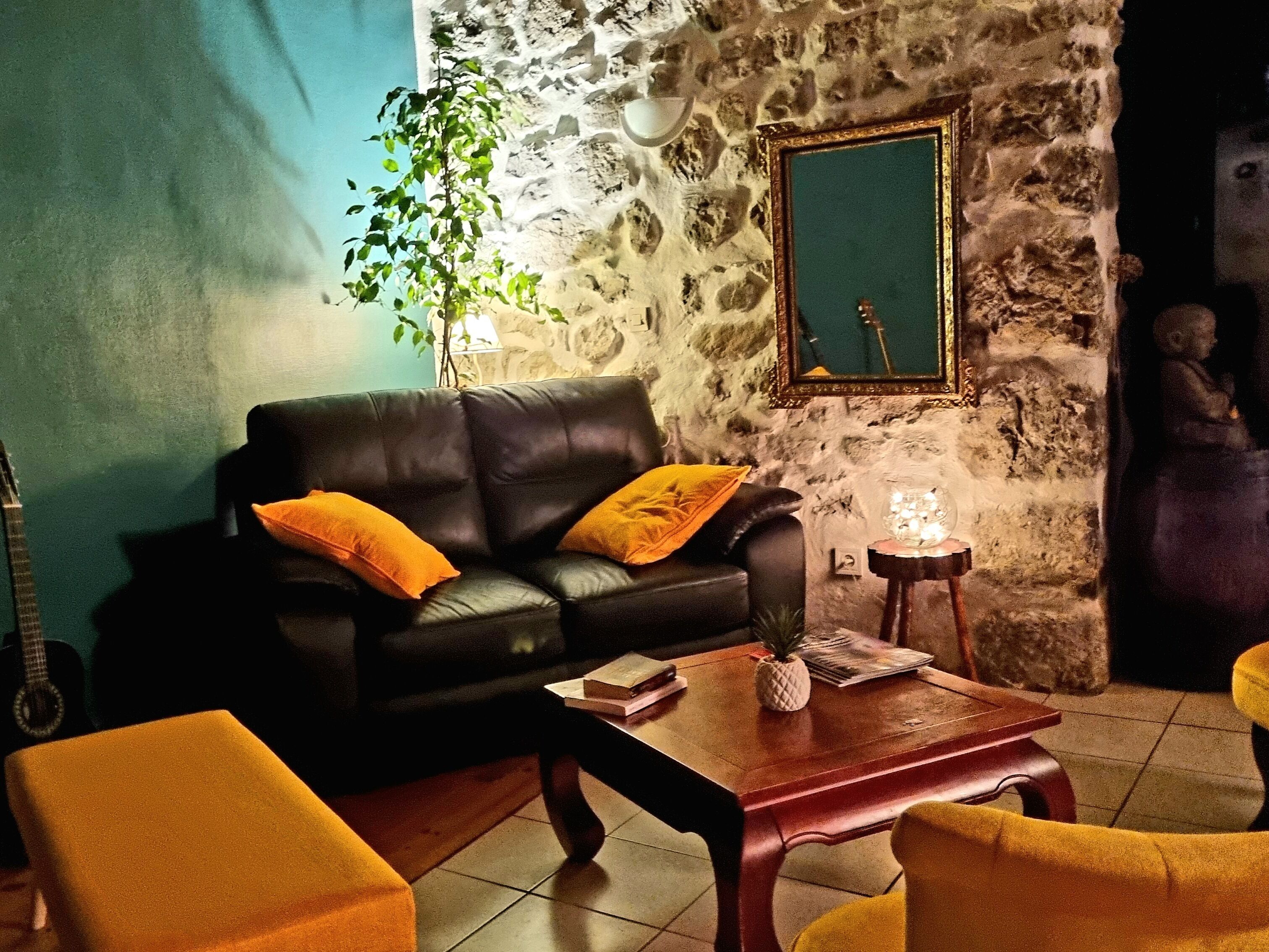 Un salon avec un canapé en cuir noir, deux coussins jaunes, une table basse en bois, une petite table d’appoint sur laquelle se trouve un ananas, et un mur en pierre agrémenté d’un miroir.