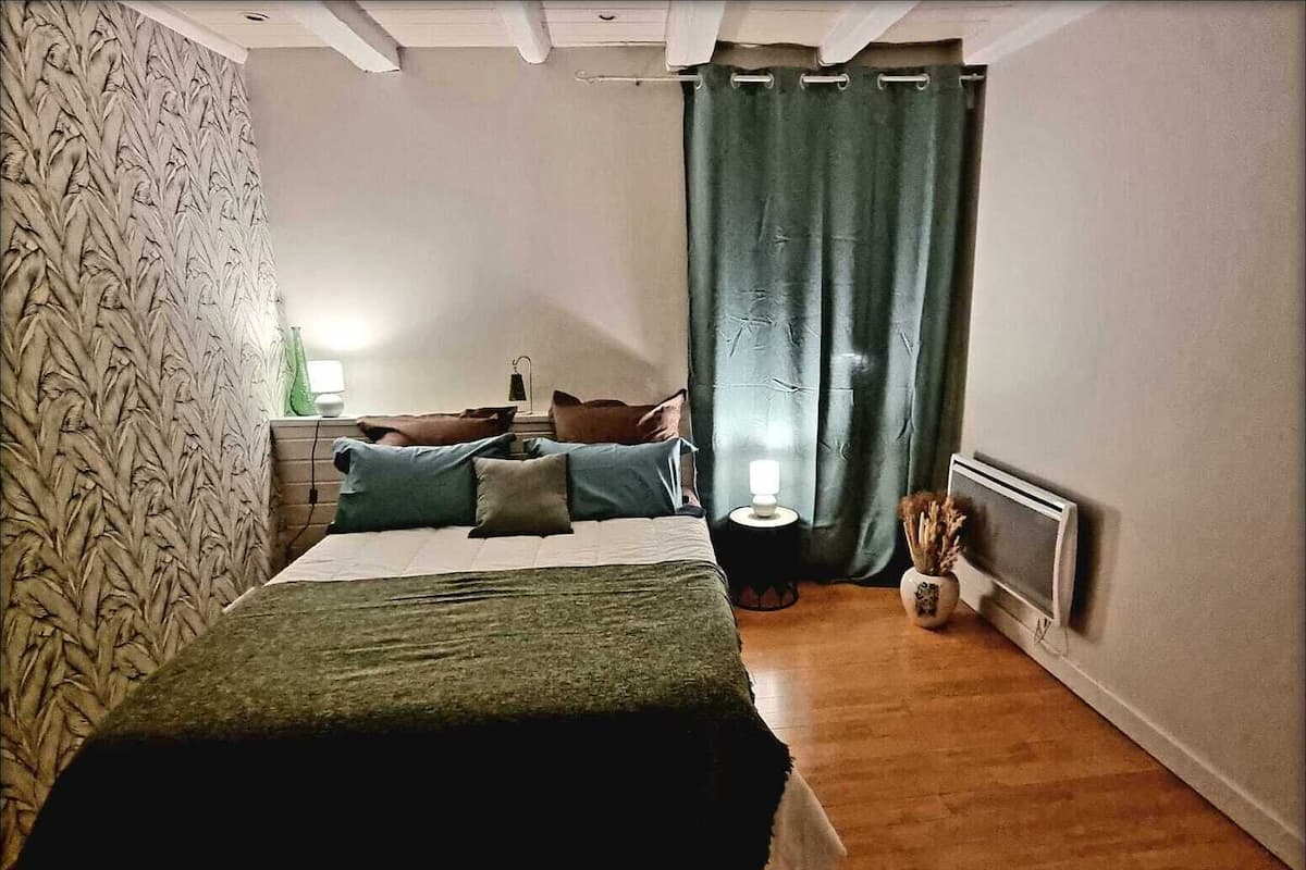 Un dormitorio con cama, mesita de noche, lámpara, radiador y cortina.