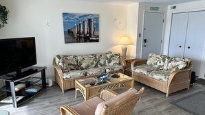 A living room with a sofa, a chair, a coffee table, a TV, and a painting of a pier. - Thunder Island 157d 2 Bedroom Condo by RedAwning (Ocean City)