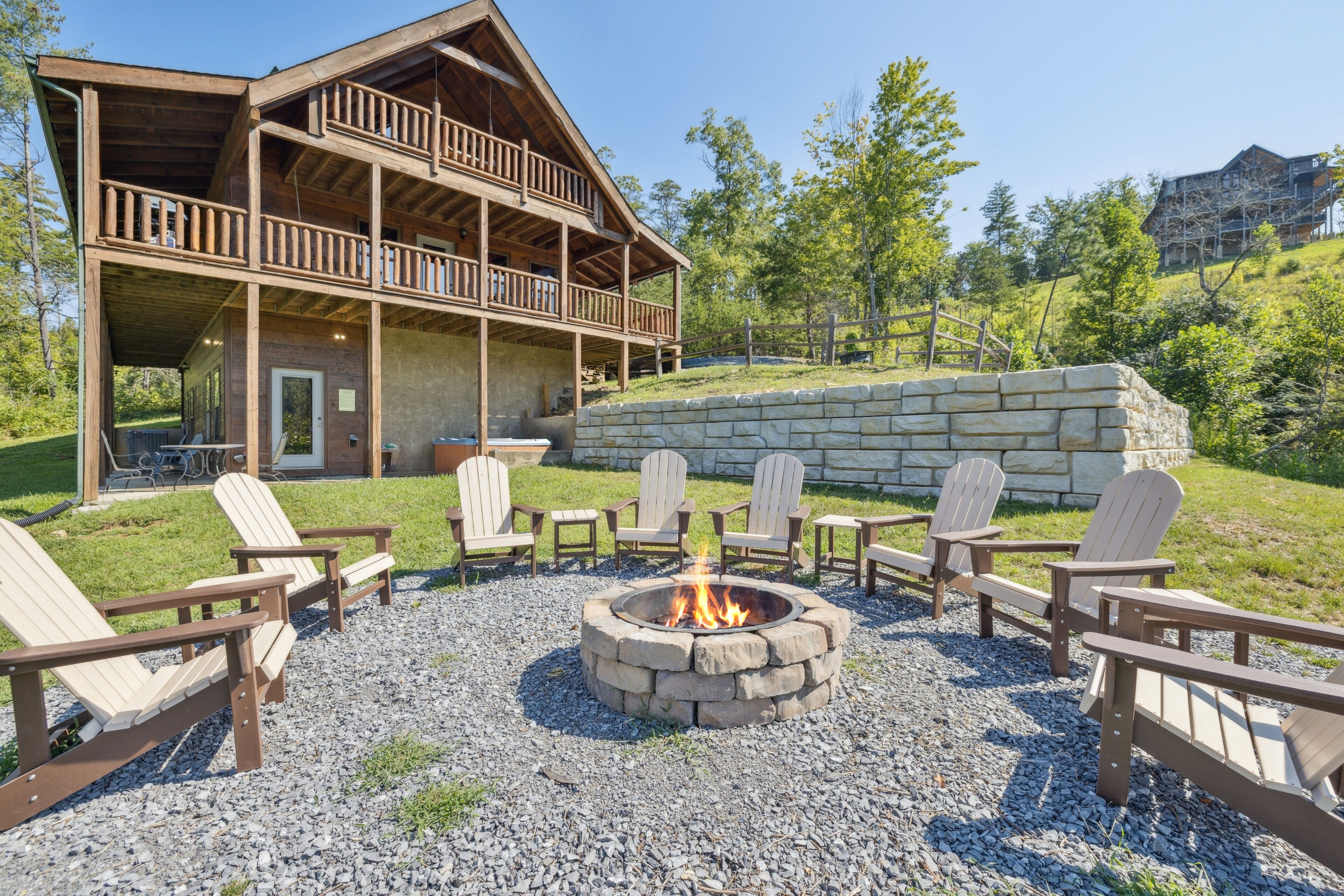 A lodge with a stone fire pit surrounded by Adirondack chairs, a covered deck, and a stone retaining wall.