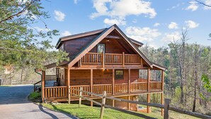 A two-story log cabin with a balcony, surrounded by a wooden fence and a forested landscape.