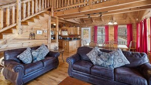 A wooden cabin interior with a staircase, two leather sofas with patterned cushions, a dining area, and a kitchen.