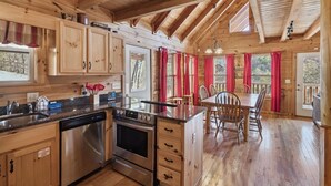 A wooden cabin with a kitchen featuring stainless steel appliances, a dining area with a table and chairs, and large windows with red curtains.