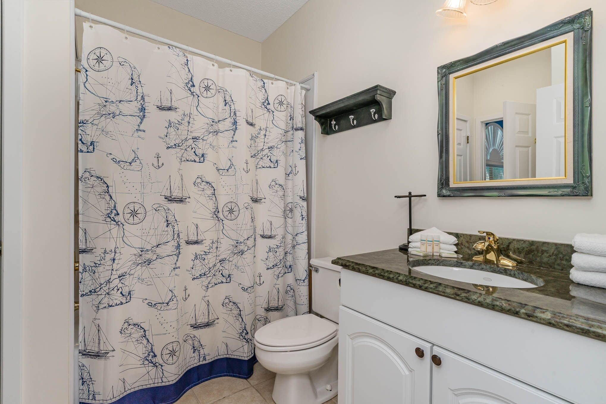 Une salle de bain avec un rideau de douche à motifs nautiques, des toilettes blanches, un meuble-lavabo avec un robinet doré et un miroir avec un cadre doré.