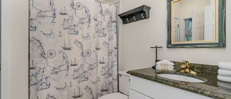 Une salle de bain avec un rideau de douche à motifs nautiques, des toilettes blanches, un meuble-lavabo avec un robinet doré et un miroir avec un cadre doré.