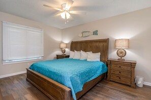 A bedroom with a wooden bed, a ceiling fan, two bedside tables with lamps, a window with blinds, and a wall decoration. - Gorgeous and spacious 4 bedroom villa - 8D Windermere by the Sea 4 Villa (Myrtle Beach)