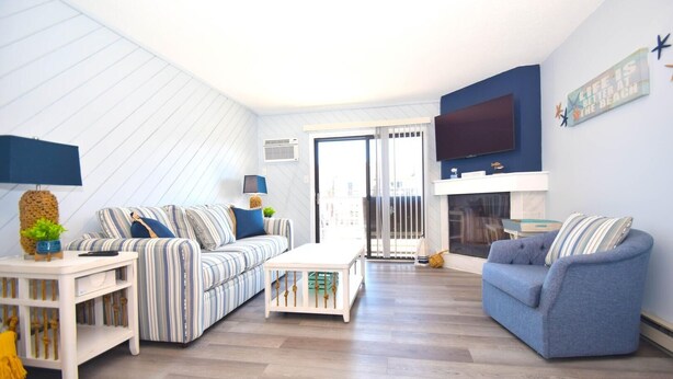 A living room with striped sofas, a coffee table, and a TV. There is a fireplace and a large window. - Harbour Bay 34 2 Bedroom Townhouse by RedAwning (Ocean City)