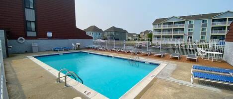 A rectangular pool with a diving board, surrounded by lounge chairs and a white fence.