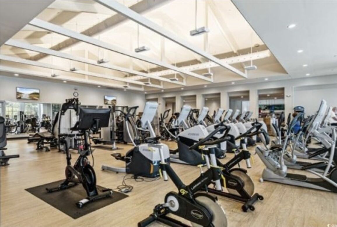 Ein großzügiger Fitnessraum mit verschiedenen Cardiogeräten, Gewichten und einer hohen Decke mit Tageslicht.