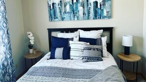 Un lit bien fait, recouvert d’une couvre-lit à motifs, un tableau représentant un paysage urbain au-dessus, et deux tables de chevet avec des lampes.