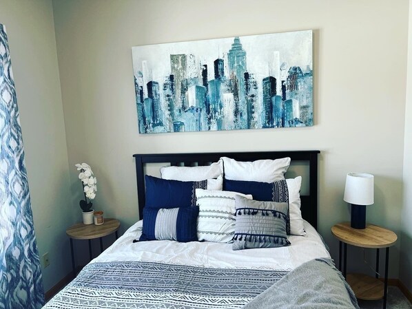 A neatly made bed with a patterned bedspread, a cityscape painting above it, and two bedside tables with lamps.