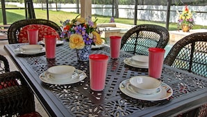 Une terrasse couverte avec une table dressée pour quatre personnes, agrémentée de chaises en osier, d’un vase de fleurs et de tasses rouges.