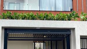 Ein modernes, zweistöckiges Gebäude mit flachem Dach, einem Balkon mit Glasgeländern und einer überdachten Terrasse mit Tisch und Stühlen.