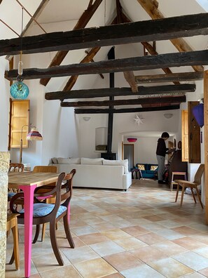 Un interior rústico con vigas de madera a la vista, una mesa de comedor con sillas, un sofá y una persona cerca de una escalera.