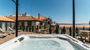A rooftop terrace with a hot tub, outdoor seating, and a view of buildings and hills.