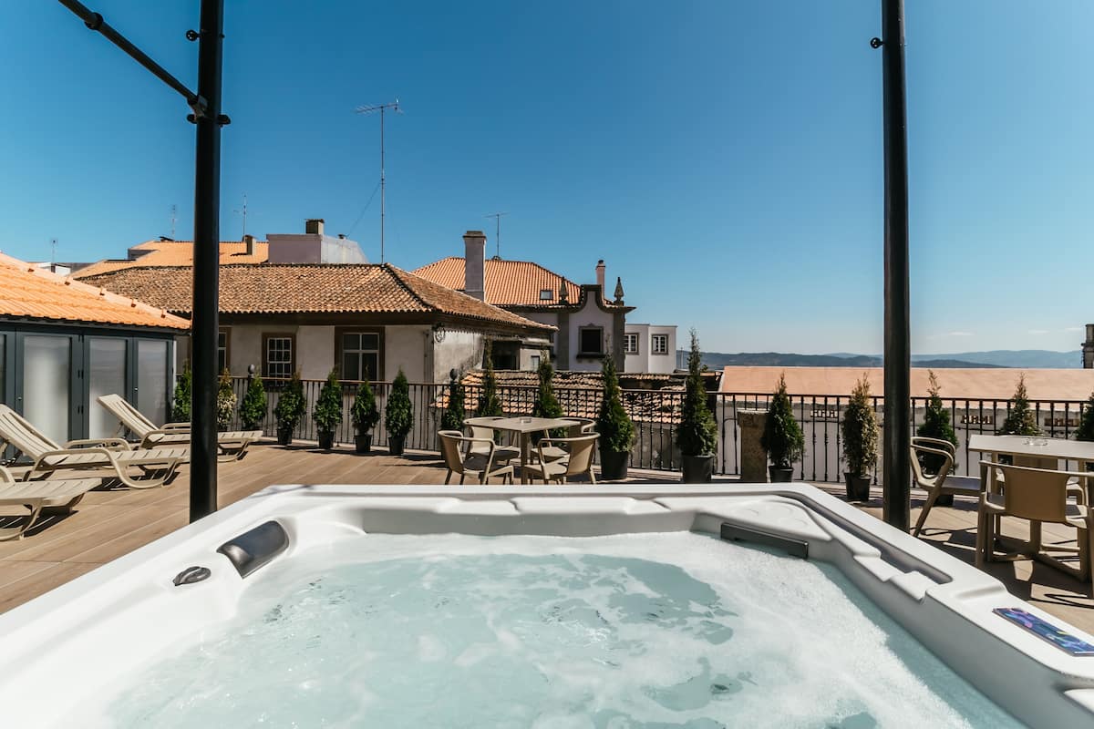 A rooftop terrace with a hot tub, outdoor seating, and a view of buildings and hills.