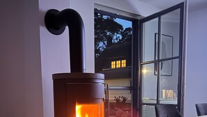 A modern living room with a wood-burning stove, a glass coffee table, a white furry chair, and a view of the outside through a glass door. - Vacation home Fischerhuus on Usedom with sauna, fireplace and private garden (Zirchow)