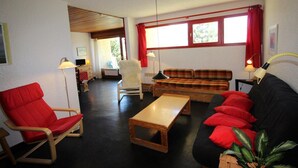 A living room with a red chair, a wooden coffee table, a black sofa with red cushions, a plaid sofa, and a white armchair.