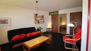 A living room with a black sofa, red cushions, a wooden coffee table, a red armchair, a floor lamp, a bookshelf, and a door leading to another room.