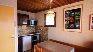 A compact kitchen with a microwave, a small refrigerator, and a tiled backsplash. There's a dining table with chairs and a poster on the wall.