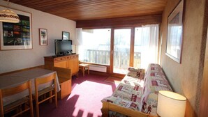 A cozy living room with a wooden ceiling, a sofa, a TV, a dining table, and a balcony with curtains.