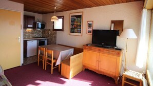 A compact kitchen with a microwave, a small refrigerator, a dining table with chairs, a wooden cabinet, a television, and a lamp.
