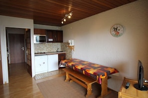 Una cocina compacta con armarios de madera, un microondas y un lavamanos. Hay un banco con una funda acolchada de colores y un televisor sobre un pedestal de madera.