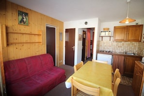 A compact living space with a red sofa, a yellow dining table, wooden cabinets, and a kitchen area including a microwave and sink.
