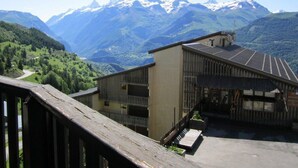 A building with a metal roof and a wooden deck, surrounded by mountains and greenery. - Ski-in/ski-out & foot of the slopes, 2nd floor, balcony, tv, 23m², Auris en Oisans (Auris)