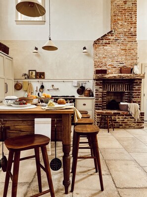 Una cocina con chimenea de ladrillo, taburetes de madera y utensilios de cocina colgando.