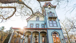 Une grande maison de style victorien, richement décorée, avec une tour imposante, une clôture blanche en bois et une véranda ornée de guirlandes.