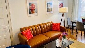 Una sala de estar con un sofá de cuero marrón, dos almohadas rojas, una mesa de centro con un jarrón de flores y cuadros en la pared.