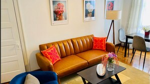 Una sala de estar con un sofá de cuero marrón, un sillón azul y una mesa de centro negra. Hay dos cuadros enmarcados en la pared, una lámpara de pie y una mesa de comedor con sillas.