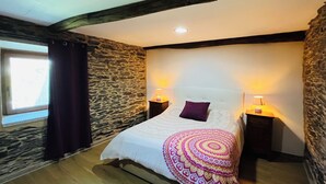 Une chambre avec un mur en pierre, des poutres apparentes, un lit recouvert d’une couverture à motifs, deux lampes de chevet et une fenêtre avec des rideaux.