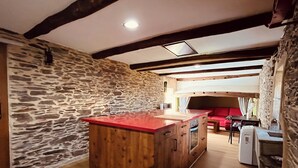 Une cuisine dotée d’un îlot central, de murs en pierre, de poutres en bois et d’un canapé rouge en arrière-plan.