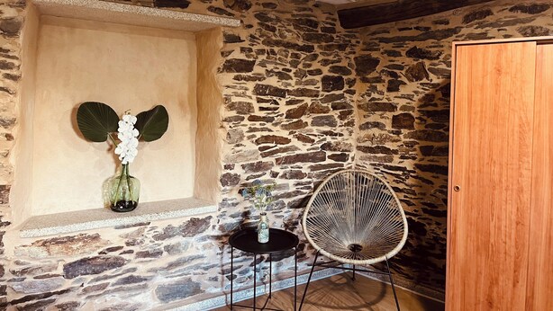 Une pièce aux murs de pierre, dotée d’une porte en bois, d’une petite table avec une chaise et d’un vase rempli de fleurs.