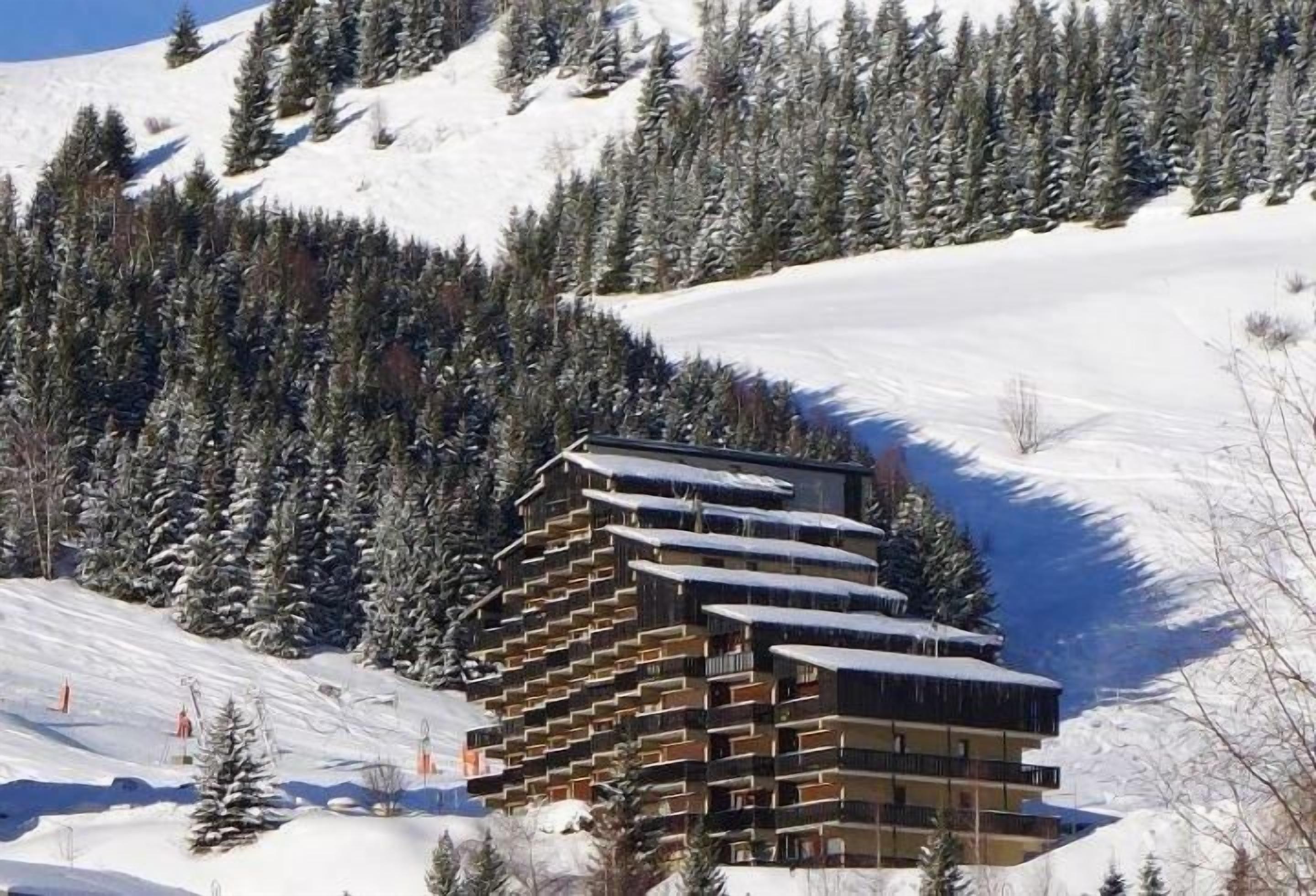 A multi-story building with balconies, surrounded by snow-covered trees and a ski slope.