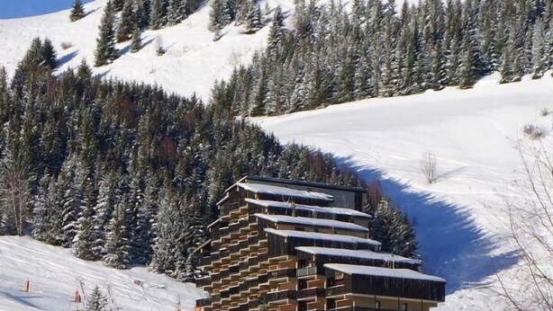 Un chalet à étages doté d’une architecture originale et superposée, entouré d’arbres couverts de neige et situé à proximité d’une piste de ski.