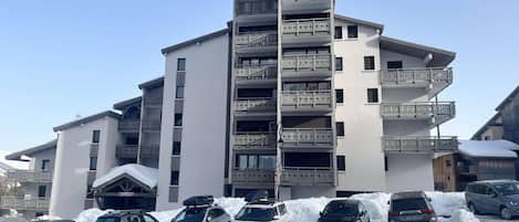 A multi-story building with balconies, a parking lot with several cars, and snow on the ground.