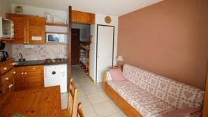 A compact kitchen with wooden cabinets, a microwave, and a small refrigerator. A dining table with chairs is positioned in front of the kitchen. A sofa with a heart-patterned cover is placed against a wall in the living area. - View mountain, balcony, parking, tv, ski locker, 23m², Auris en Oisans (Auris)