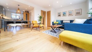 A modern living room with a blue sofa, yellow armchair, and a dining area with a wooden table and chairs. - Huckleberry 4-Bed Home (Newquay)