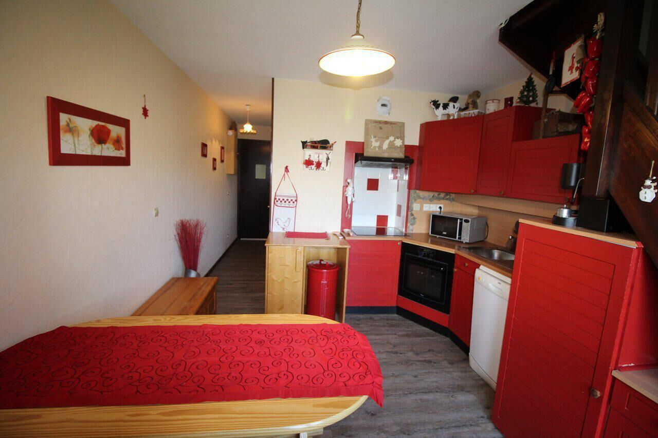 A compact room with a bed, a small table, a kitchenette with red cabinets, and a wooden bench.