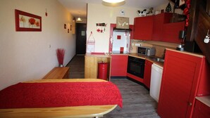 Une pièce de taille modeste comprenant un lit, une petite table, un coin cuisine avec des placards rouges et un banc en bois.