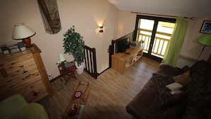 Un salon chaleureux avec un plancher en bois, un canapé marron, une télévision, une plante et un escalier menant à un balcon.