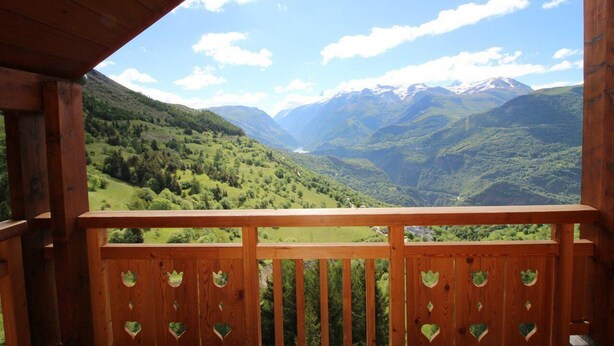 A wooden balcony with heart-shaped cutouts, overlooking a valley with green hills and distant snow-capped mountains under a clear sky.