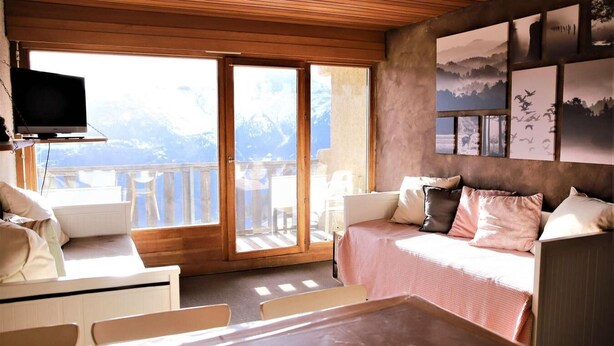 A cozy room with a bed, a table, and a TV. It has a balcony with a view of mountains.