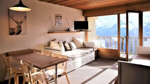 A cozy room with a wooden ceiling, a sofa with cushions, a dining table with chairs, and a large window offering a mountain view.