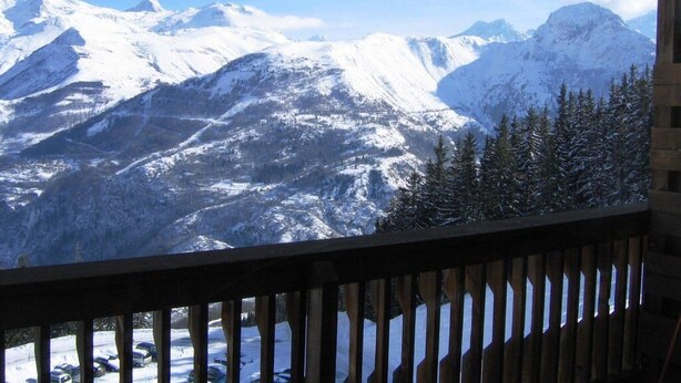 A mountain view from a wooden balcony with snow-covered peaks and pine trees.