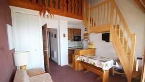 A cozy room with a bed, a wooden bench, a table with a tablecloth, a wooden staircase, and a kitchen area with cabinets and appliances.